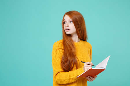 Young redhead woman girl in yellow knitted sweater posing isolated on blue turquoise background studio portrait. People lifestyle concept. Mock up copy space. Writing notes in notebook, looking aside.の写真素材