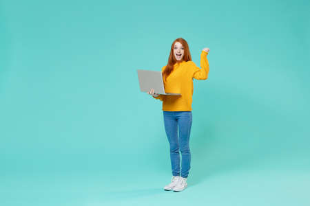 Excited young redhead girl in yellow sweater posing isolated on blue turquoise wall background in studio. People lifestyle concept. Mock up copy space. Work on laptop pc computer doing winner gesture.の写真素材