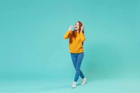 Funny young redhead girl in yellow knitted sweater posing isolated on blue turquoise background studio portrait. People lifestyle concept. Mock up copy space. Listen music with headphones, sing song.の写真素材