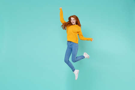 Funny young redhead woman girl in yellow knitted sweater posing isolated on blue turquoise background studio portrait. People emotions lifestyle concept. Mock up copy space. Jumping, spreading hands.の写真素材