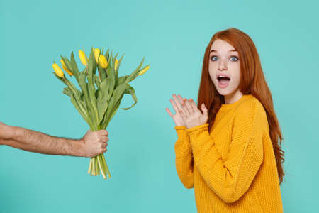 Surprised young redhead woman girl in yellow sweater posing isolated on blue turquoise background in studio. People lifestyle concept. Mock up copy space. Taking bouquet of yellow tulips in male hand.の写真素材