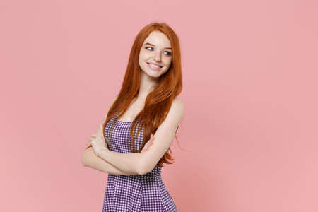 Smiling young redhead woman girl in plaid dress posing isolated on pastel pink background studio portrait. People emotions lifestyle concept. Mock up copy space. Holding hands crossed, looking aside.の写真素材