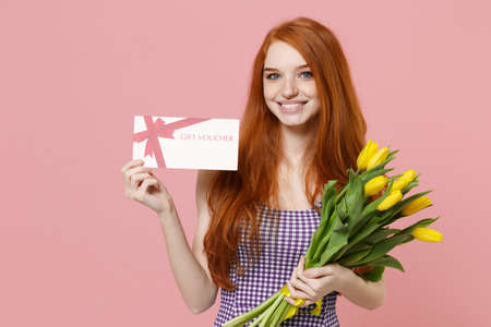 Smiling young redhead woman girl in plaid dress posing isolated on pastel pink wall background in studio. People lifestyle concept. Mock up copy space. Hold bouquet of yellow tulips, gift certificate.の写真素材