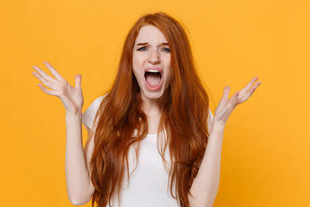 Frustrated young redhead woman girl in white blank empty t-shirt posing isolated on yellow background studio portrait. People emotions lifestyle concept. Mock up copy space. Spreading hands screaming.の写真素材
