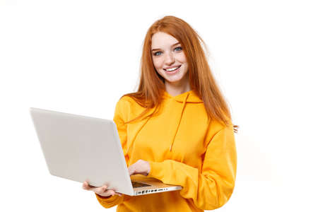 Smiling young redhead woman girl in yellow hoodie posing isolated on white background studio portrait. People sincere emotions lifestyle concept. Mock up copy space. Working on laptop pc computer.の写真素材