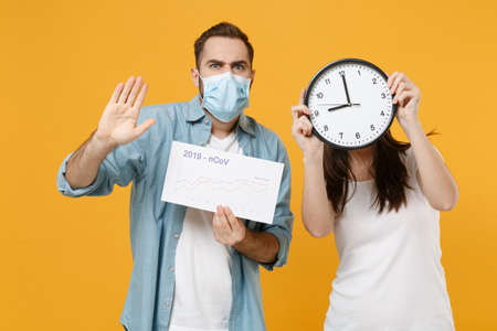 Two shocked people in sterile face masks hold clock isolated on yellow background studio. Epidemic...