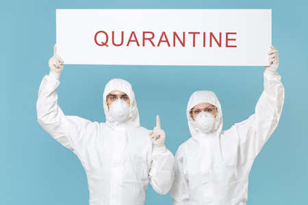 People in protective suit respirator mask hold big white with text quarantine sign board isolated on blue background studio. Epidemic rapidly spreading coronavirus 2019-ncov medicine flu virus conceptの写真素材