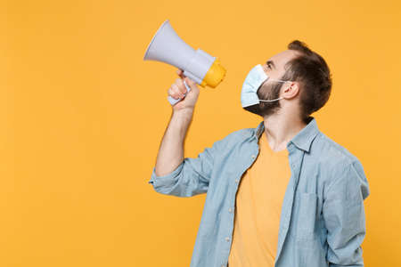 Side view of young man in sterile face mask posing isolated on yellow wall background studio portrait. Epidemic pandemic coronavirus 2019-ncov sars covid-19 flu virus concept. Scream in megaphone.の写真素材
