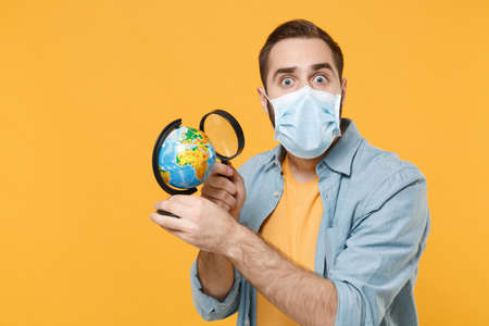 Shocked young man in sterile face mask posing isolated on yellow background studio portrait. Epidemic pandemic coronavirus 2019-ncov sars covid-19 flu virus concept. Hold magnifying glass world globe.の写真素材