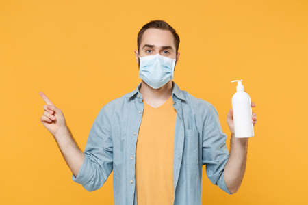 Young man in face mask isolated on yellow background. Epidemic pandemic coronavirus 2019-ncov sars covid-19 flu virus concept. Hold bottle antibacterial sanitizer soap pointing index finger aside up.の写真素材