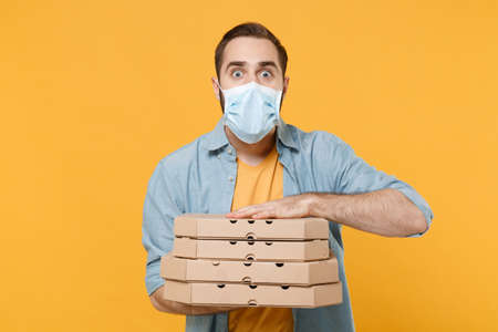 Shocked young man in sterile face mask posing isolated on yellow background in studio. Epidemic pandemic coronavirus 2019-ncov sars covid-19 flu virus concept. Hold italian pizza in cardboard flatbox.の写真素材
