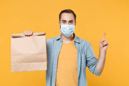 Young man in face mask isolated on yellow background. Epidemic pandemic coronavirus 2019-ncov sars covid-19 flu virus concept. Hold brown empty blank craft paper bag for takeaway pointing finger up.の写真素材