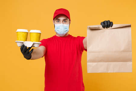 Delivery man guy employee in red cap uniform mask gloves hold craft paper packet with food coffee...