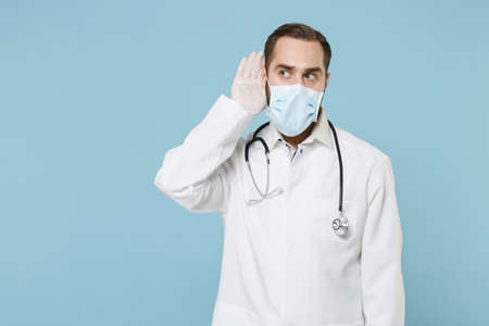 Male doctor man in white medical gown sterile face mask gloves isolated on blue background. Epidemic pandemic coronavirus 2019-ncov sars covid-19 flu virus concept. Try to hear you with hand near ear.の写真素材