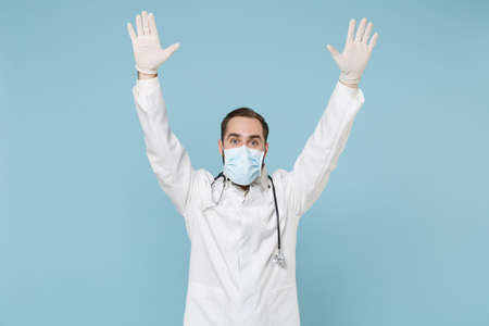 Excited male doctor man in white medical gown sterile face mask gloves isolated on blue background. Epidemic pandemic spreading coronavirus 2019-ncov sars covid-19 flu virus concept. Rising hands up.の写真素材