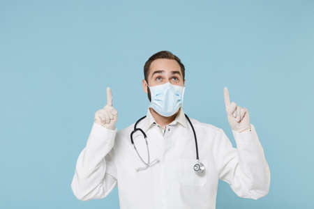 Male doctor man in white medical gown sterile face mask gloves isolated on blue background. Epidemic...