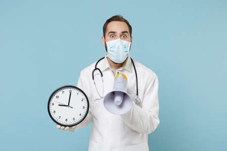 Excited male doctor man in medical gown sterile face mask gloves isolated on blue background. Epidemic pandemic coronavirus 2019-ncov sars covid-19 flu virus concept. Scream in megaphone hold clock.の写真素材