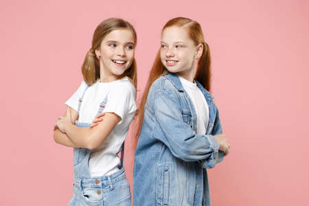 Funny little kids girls 12-13 years old in denim clothes isolated on pink background. Childhood lifestyle concept. Mock up copy space. Standing back to back, looking at each other, hold hands crossed.の写真素材