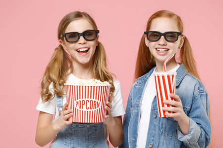 Cheerful little kids girls 12-13 years old in 3d glasses denim clothes isolated on pink background. Childhood lifestyle concept. Mock up copy space. Watching movie film hold bucket popcorn cup soda.の写真素材