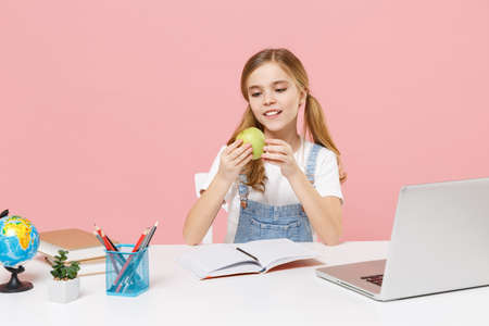 Little kid schoolgirl 12-13 years old in white t-shirt sit studying at white desk with pc laptop isolated on pink background. School distance education at home during quarantine concept. Hold apple.の写真素材