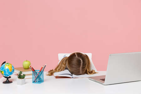 Tired little kid schoolgirl 12-13 years old sit study at desk with pc laptop isolated on pink...