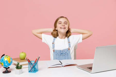 Smiling little kid schoolgirl 12-13 years old study at white desk with laptop isolated on pink background. School distance education at home during quarantine concept. Sleeping with hands behind head.の写真素材