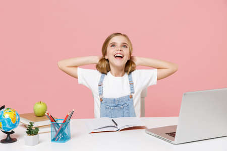 Funny little kid schoolgirl 12-13 years old study at white desk with laptop isolated on pink...