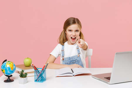 Dissatisfied little kid schoolgirl 12-13 years old study at white desk with pc laptop isolated on pink background. School distance education at home concept. Pointing index finger on camera screaming.の写真素材