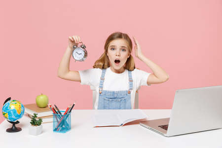 Shocked worried little kid schoolgirl 12-13 years old sit study at white desk with pc laptop isolated on pink background. School distance education at home during quarantine concept. Hold alarm clock.の写真素材
