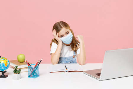 Exhausted little kid schoolgirl 12-13 years old in face mask sit study at desk with laptop isolated...