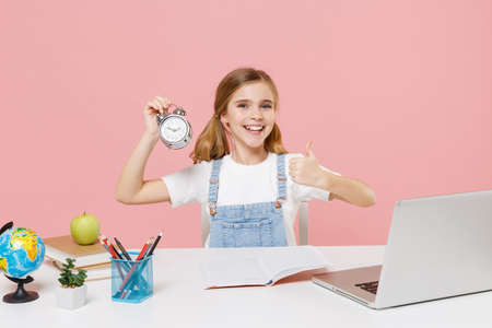 Smiling little kid schoolgirl 12-13 years old sit study at desk with laptop isolated on pink background. School distance education at home during quarantine concept. Hold alarm clock showing thumb up.の写真素材