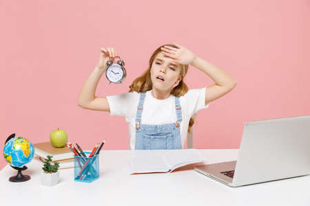 Tired little kid schoolgirl 12-13 years old sit study at desk with laptop isolated on pink...