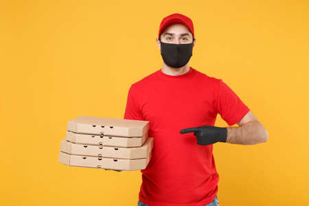 Delivery man employee in red cap t-shirt uniform black mask gloves give food order pizza boxes isolated on yellow background studio. Service quarantine pandemic coronavirus virus flu 2019-ncov conceptの写真素材