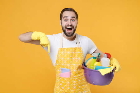 Amazed young man househusband in apron rubber gloves hold basin with detergent bottles washing cleansers doing housework isolated on yellow background. Housekeeping concept. Point index finger down.の写真素材
