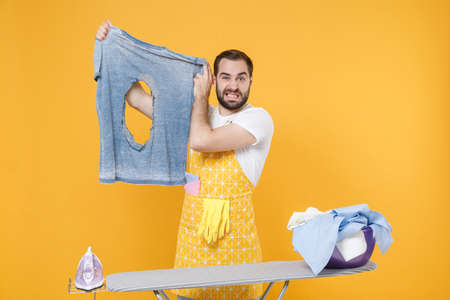 Confused young man househusband in apron ironing clean clothes on board while doing housework isolated on yellow wall background studio. Housekeeping concept. Mock up copy space. Hold burnt t-shirt.の写真素材