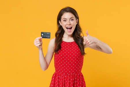 Excited young brunette woman girl in red summer dress posing isolated on yellow wall background studio portrait. People lifestyle concept. Mock up copy space. Hold credit bank card, showing thumb up.の写真素材