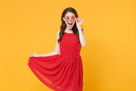Excited young brunette woman girl in red summer dress, eyeglasses posing isolated on yellow wall...