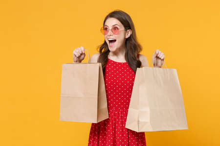 Surprised young brunette woman girl in red summer dress, eyeglasses posing isolated on yellow wall background studio portrait. People lifestyle concept. Hold package bag with purchases after shopping.の写真素材