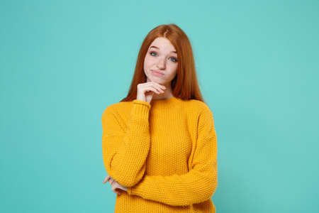 Confused puzzled young redhead woman girl in yellow sweater posing isolated on blue turquoise wall background studio portrait. People lifestyle concept. Mock up copy space. Put hand prop up on chin.の写真素材