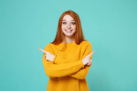 Excited young redhead woman girl in yellow sweater posing isolated on blue turquoise wall background. People lifestyle concept. Mock up copy space. Pointing index fingers aside, holding hands crossed.の写真素材