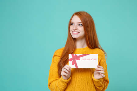 Smiling young redhead woman girl in yellow knitted sweater posing isolated on blue turquoise background in studio. People lifestyle concept. Mock up copy space. Hold gift certificate looking aside.の写真素材