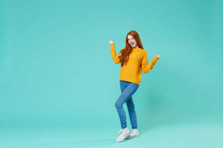 Happy young redhead woman girl in yellow sweater posing isolated on blue turquoise wall background studio portrait. People sincere emotions lifestyle concept. Mock up copy space. Doing winner gesture.の写真素材