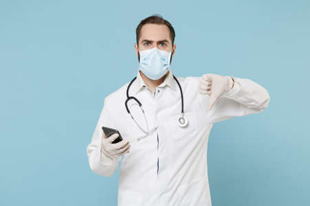 Male on-line doctor man in medical gown face mask gloves isolated on blue background. Epidemic pandemic coronavirus 2019-ncov sars covid-19 flu virus concept. Using mobile phone showing thumb down.の写真素材