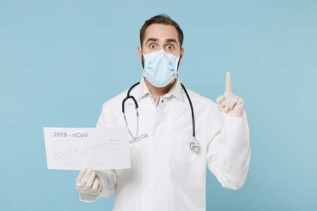Doctor man in medical gown face mask gloves isolated on blue background. Epidemic pandemic coronavirus 2019-ncov sars covid-19 flu virus concept. Hold card with globe infographiÑ, point finger up.の写真素材