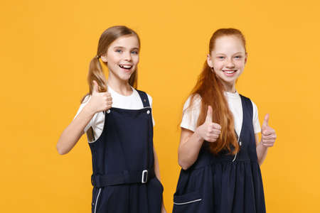 Two young little girls 12-13 years old in white t-shirt blue school uniform dresses isolated on yellow background children studio portrait Childhood kids education lifestyle concept Mock up copy spaceの写真素材