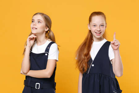Two young little girls 12-13 years old in white t-shirt blue school uniform dresses isolated on yellow background children studio portrait Childhood kids education lifestyle concept Mock up copy spaceの写真素材