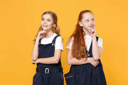 Two young little girls 12-13 years old in white t-shirt blue school uniform dresses isolated on yellow background children studio portrait Childhood kids education lifestyle concept Mock up copy spaceの写真素材