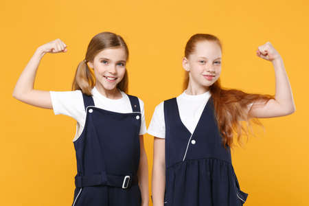 Two young little girls 12-13 years old in white t-shirt blue school uniform dresses isolated on yellow background children studio portrait Childhood kids education lifestyle concept Mock up copy spaceの写真素材