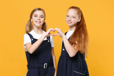 Two young little girls 12-13 years old in white t-shirt blue school uniform dresses isolated on yellow background children studio portrait Childhood kids education lifestyle concept Mock up copy spaceの写真素材