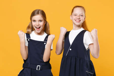 Two young little girls 12-13 years old in white t-shirt blue school uniform dresses isolated on yellow background children studio portrait Childhood kids education lifestyle concept Mock up copy spaceの写真素材